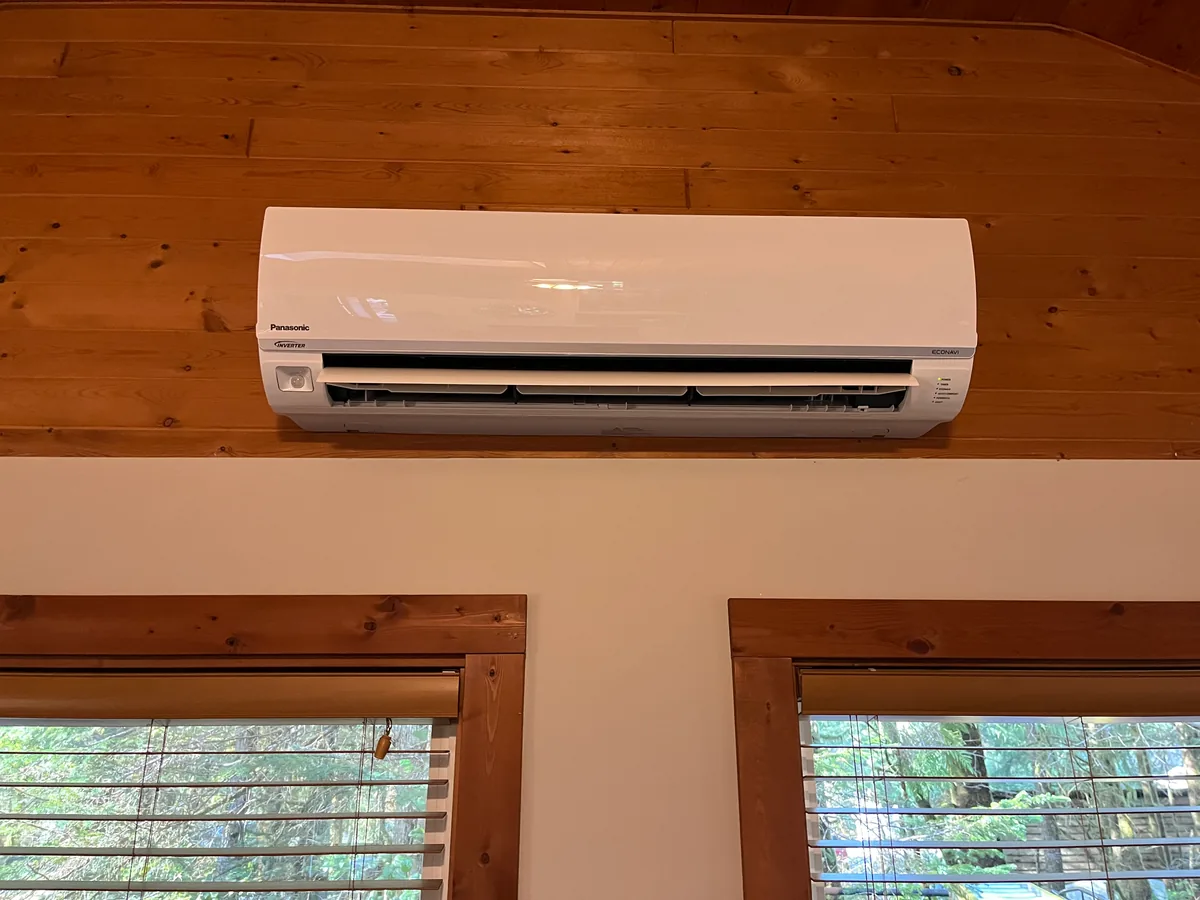 A ductless heat pump head mounted high on a wood-panelled wall above two windows