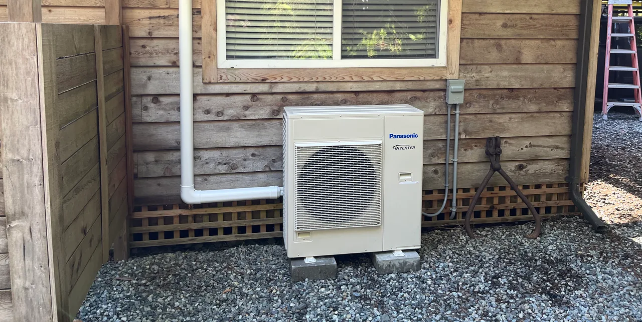 A ductless heat pump outdoor unit on concrete blocks beside cedar siding, with a white line set cover running up the wall