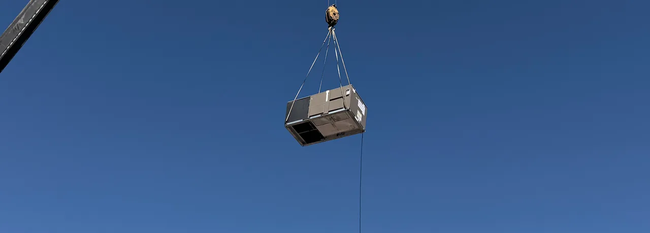 A package rooftop unit hanging from a crane hook against a clear blue sky