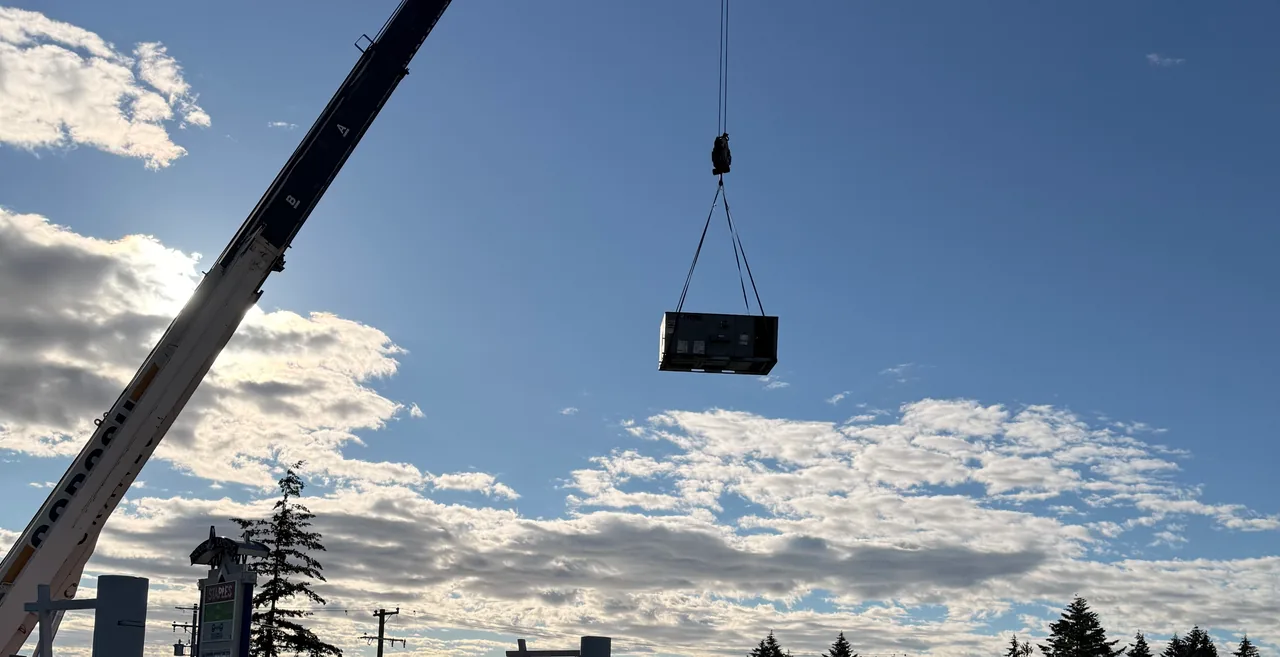 A rooftop unit on a crane hook above a flat roof, with the crane boom against a low sun