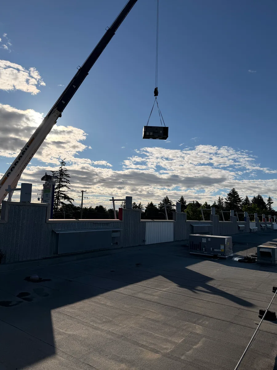 A crane lifting a rooftop unit above a flat commercial roof against a low sun