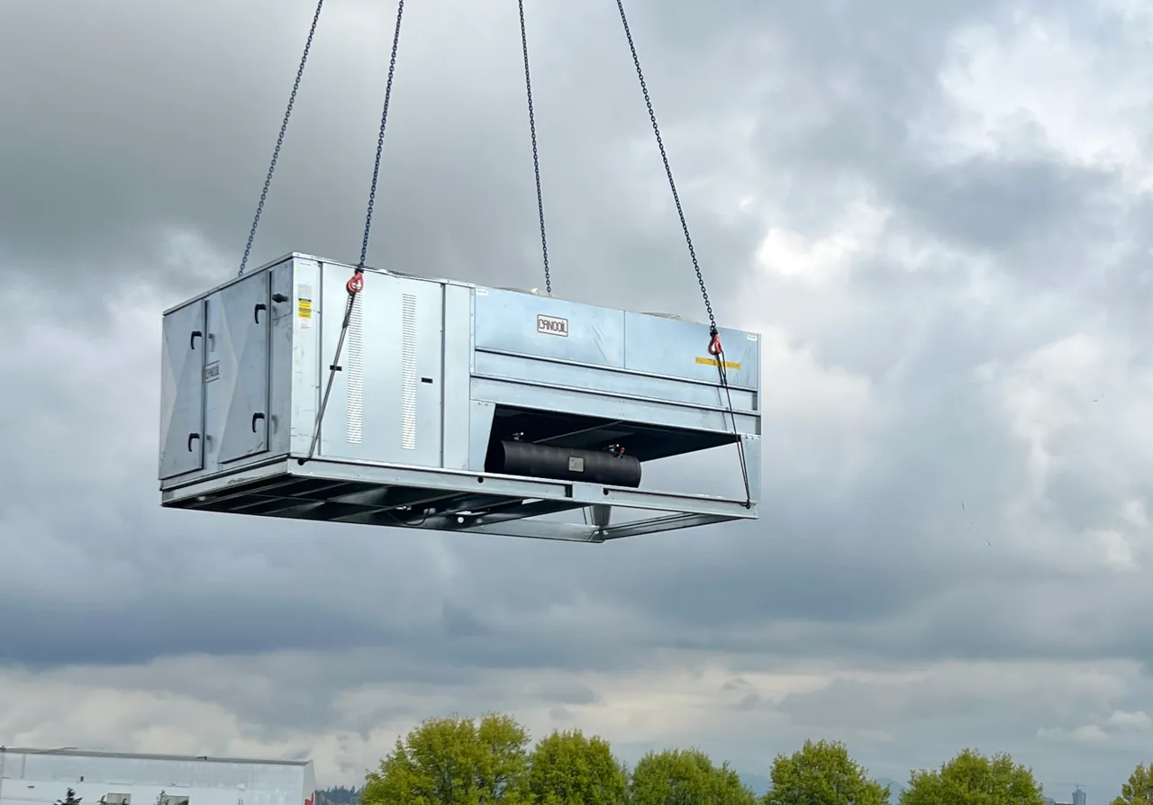 A refrigeration unit lifted on crane chains above a flat roof under a cloudy sky