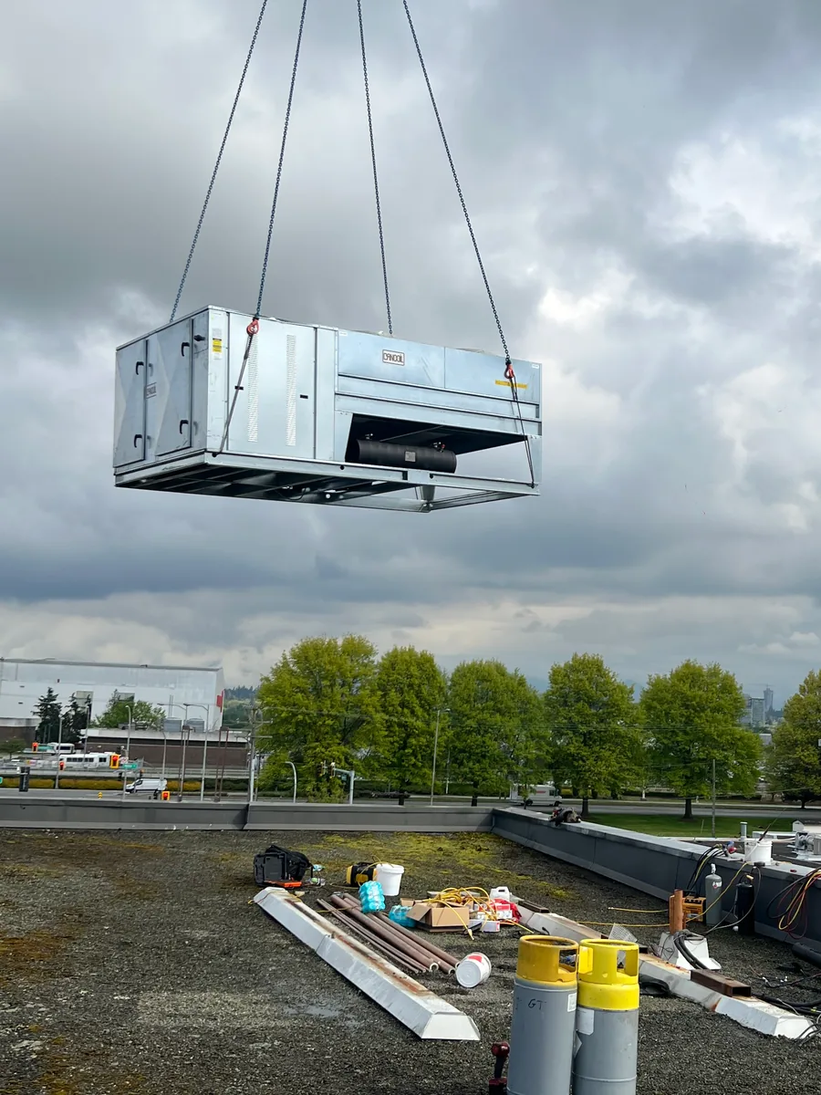A refrigeration unit lifted on crane chains above a flat roof, with refrigerant cylinders and copper pipe on the roof below