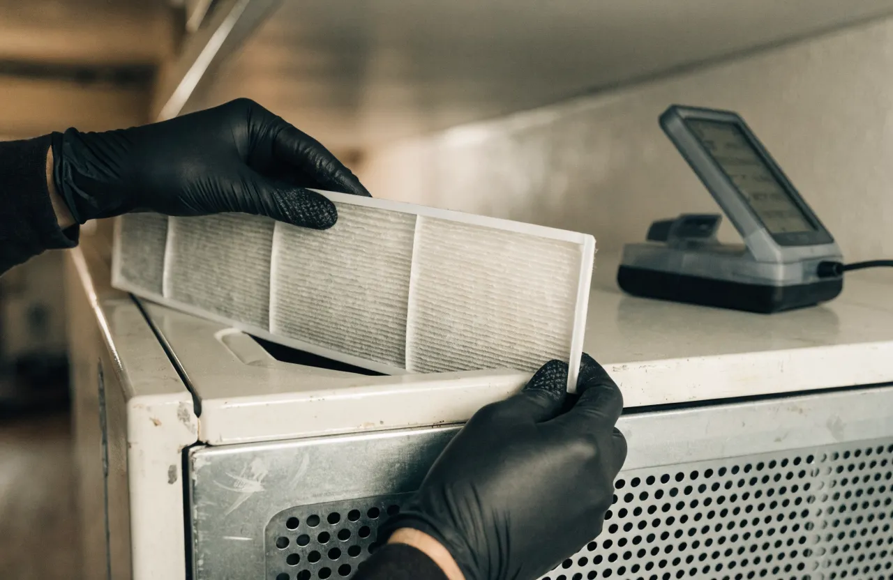 Gloved hands lifting a used grey pleated filter out of a unit, with a digital meter beside it