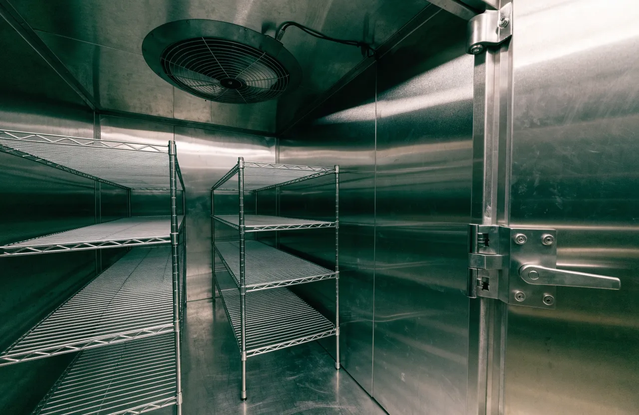 Inside a stainless steel walk-in cooler: wire shelving, a ceiling evaporator fan and a heavy latch